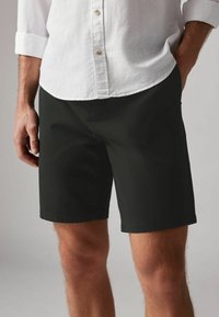 Homme portant un short noir jusqu'aux genoux et une chemise blanche à manches longues boutonnée avec les manches retroussées, se tenant les mains dans les poches.
