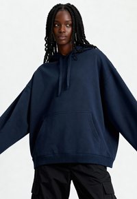 Felpa oversized blu navy in tessuto morbido con tasca frontale, cappuccio con coulisse e polsini e orlo a coste. Indossata con pantaloni cargo neri.
