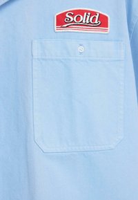 Ανοιχτό μπλε πουκάμισο από τζιν ύφασμα, με κόκκινο και λευκό έμβλημα "Solid" και τσέπη με πατς στην αριστερή πλευρά.