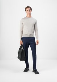 Mann in beigem Pullover, mit blauen Hosen und schwarzen Schuhen, der eine schwarze Ledertasche hält und vor einem weißen Hintergrund steht.