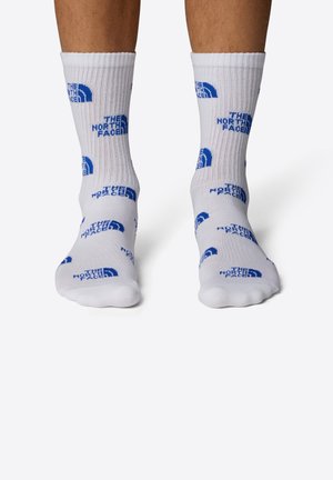 Chaussettes blanches avec texture côtelée et logo imprimé bleu. Présente "THE NORTH FACE" et un design circulaire. Visibles au-dessus de la cheville.