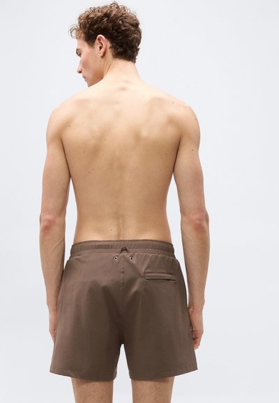 Νεαρός άνδρας με σγουρά μαλλιά, γυρισμένος πλάτη, φοράει καφέ μαγιό με πίσω τσέπη και οπές κοντά στη μέση.