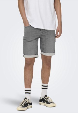 Man met opgerolde grijze denimshorts, wit T-shirt, witte sneakersokken met zwarte strepen en zwarte lage sneakers.