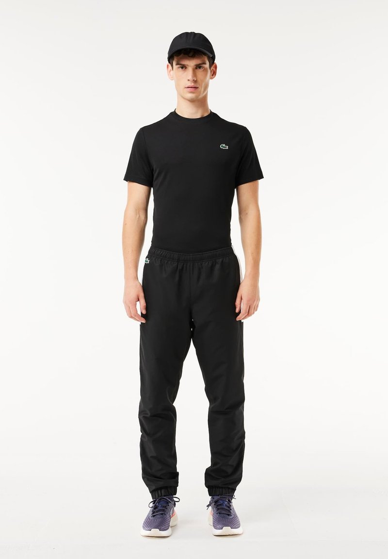 Tenue de sport noire comprenant un t-shirt ajusté à manches courtes et un pantalon à taille élastique. Le modèle porte une casquette noire avec des baskets violettes.