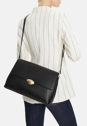 Mujer con blazer blanco de rayas finas y vaqueros oscuros, llevando un bolso de hombro de cuero negro con cierre dorado.