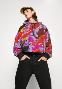 Capuchon sweatshirt in levendig rood, paars en zwart met sierlijke patronen. Heeft een relaxte pasvorm, contrasterende boorden en een verstelbare trekkoord.