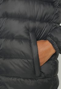 Hand delvis inskjuten i sidofickan på en svart, vadderad jacka med elastisk mudd på ärmen.