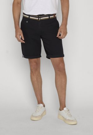 Man draagt zwarte opgerolde korte broek, een gestreepte riem, witte sneakers en een wit t-shirt, staand tegen een effen achtergrond.