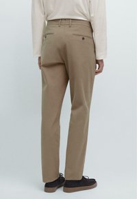 Beige Baumwollhosen mit bequemer Passform, die zwei Gesäßtaschen und einen Gürtelschlaufenbund aufweisen, kombiniert mit dunklen Schuhen.