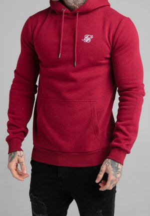 Mand iført en tætsiddende rød hættetrøje med snore og frontlomme, kombineret med revnede sorte jeans, der viser tatoveringer på hænder og hals.