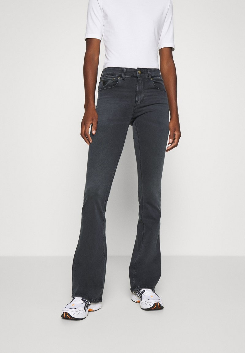 Personne portant un jean évasé gris foncé ajusté, un haut blanc à manches courtes et des baskets blanches avec des détails noirs et orange, debout sur un sol gris.
