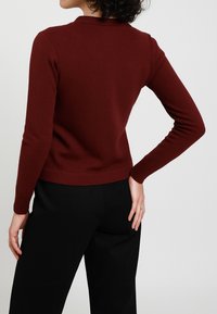 Maglione a maglia bordeaux con colletto e polsini a costine, design attillato e texture liscia, abbinato a pantaloni neri.