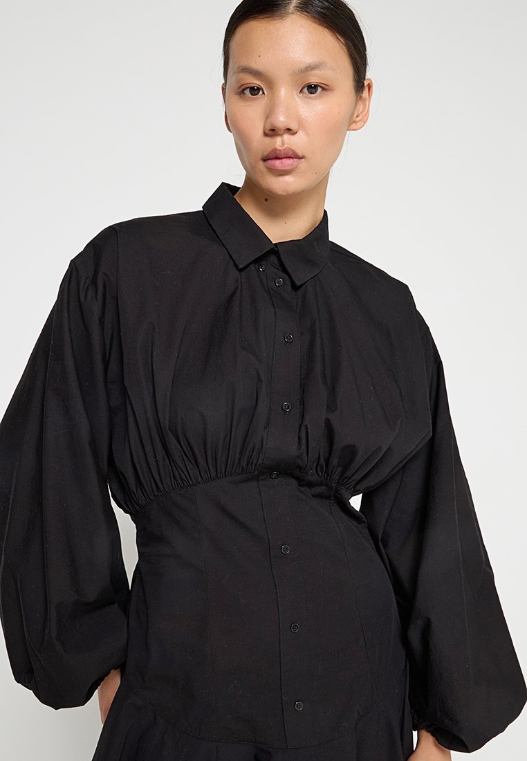 Junge Frau mit dunklem Haar, das zurückgebunden ist, trägt ein schwarzes langärmliges Hemdblusenkleid mit geraffter Taille und Puffärmeln.