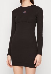 Black long-sleeve ribbed dress with a fitted silhouette, featuring a rounded neckline and a small logo patch on the upper chest.