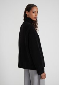 Femme aux cheveux bouclés portant un pull à col roulé noir et un pantalon gris clair, debout de profil avec une expression neutre.