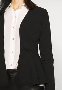 Frau trägt eine weiße Bluse mit Knöpfen unter einem schwarzen Peplum-Blazer mit langen Ärmeln vor einem einfarbigen Hintergrund.