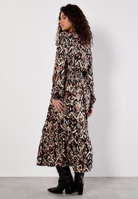 Maxikleid mit einem Muster aus Schwarz, Creme und Rosen, ausgestattet mit langen Ärmeln, einer smockierten Taille und gestuften Rüschen am Saum, kombiniert mit schwarzen Stiefeln.