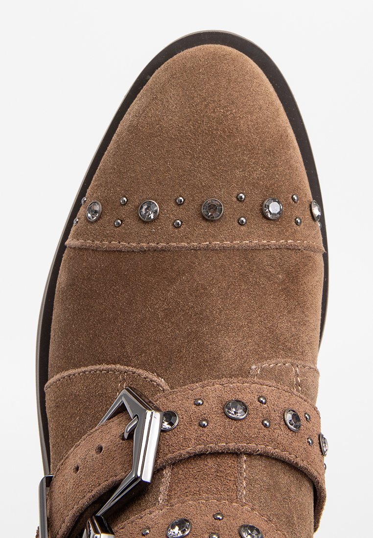 Scarpa in suede marrone con punta a punta, caratterizzata da borchie decorative e una fascia con fibbia metallica. Superficie texturizzata con una finitura liscia.