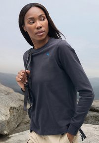 Langarm, donkergrijze shirt van zacht materiaal, met een blauw driehoeklogo op de linkermouw en een relaxte pasvorm, gecombineerd met beige broek.