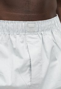 Κοντινή λήψη της λευκής ζώνης της εσώρουχης boxer Calvin Klein που φοριέται από άτομο με σκοτεινό τόνο δέρματος.