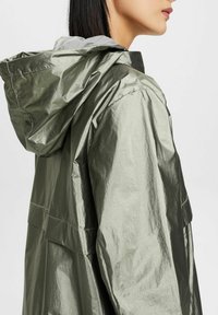 Oliwkowo-zielona metaliczna kurtka z kapturem o gładkiej, błyszczącej teksturze. Posiada luźny krój, dwie boczne kieszenie oraz kontrastową szaro-skalną podszewkę.