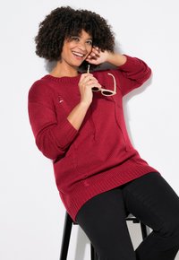 Roter Strickpullover mit lockerer Passform, geripptem Muster und überschnittenen Schultern. Getragen mit schwarzen Hosen, die einen runden Ausschnitt und Seitenschlitze aufweisen.