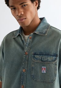 Νεαρός άνδρας που φοράει μπλε κοντομάνικο πουκάμισο από τζιν με κουμπιά μπροστά και μια μεγάλη τσέπη στο στήθος με το λογότυπο της Wrangler.