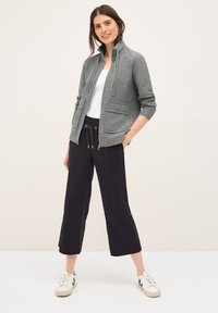 Uśmiechnięta kobieta ma na sobie szary rozpinany jacket na białej bluzce, czarne z krótkimi nogawkami spodnie i białe trampki, w tle jasne.