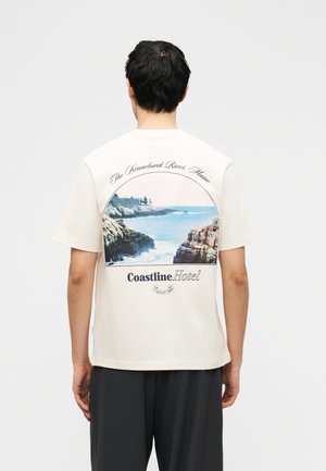 Uomo di spalle che indossa una maglietta bianca con un'immagine di scogliere costiere e oceano, accompagnata dal testo "The Kennebunk River, Maine" e "Coastline Hotel."