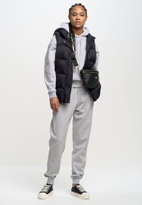 Młoda osoba ubrana w szarą bluzę z kapturem, dopasowane spodnie dresowe, czarną pikowaną kamizelkę, czarną torbę na ramię i czarne trampki z białymi podeszwami i sznurówkami.