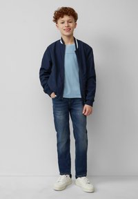 Marine bomberjack met een witte gestreepte kraag, lichtblauw T-shirt, blauwe spijkerbroek en witte sneakers. Staande pose op een neutrale achtergrond.