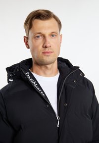 Sort hættetrøje med lynlås og høj krave, med et logo på lynlåsområdet, lavet af glat, vandafvisende materiale.
