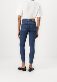 Džíny s vysokým pasem vyrobené z modrého denimu, se slim fit střihem, zadními kapsami a hnědou koženou nášivkou na pasu. V kombinaci s bílým tričkem.