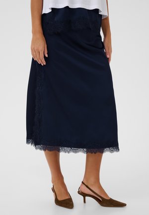 Žena s národnou midi sukňou s čipkovým lemom a hnedými semišovými slingback podpätkami, stojaca na jednoduchom svetlom pozadí.