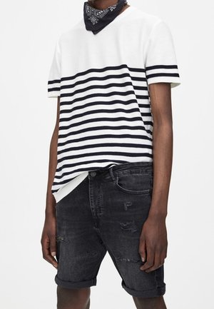 Person iført en hvid og sort stribet kortærmet skjorte, sort bandana omkring halsen og opbukket sort denimshorts med slidte detaljer.