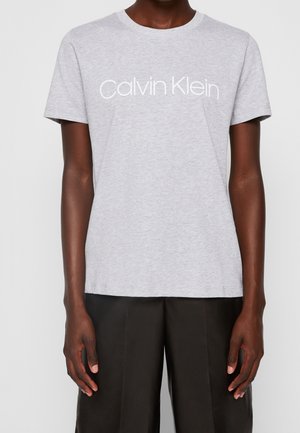 Person trägt ein hellgraues Calvin Klein T-Shirt und schwarze Hose, gezeigt von den Schultern bis zur Mitte der Oberschenkel, stehend vor schlichtem Hintergrund.