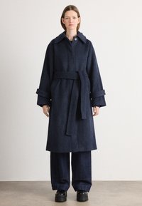 Cappotto di lana blu navy con ampio colletto, vita con cintura e maniche larghe. Presenta dettagli con bottoni e una vestibilità morbida, abbinato a pantaloni in denim a gamba larga.