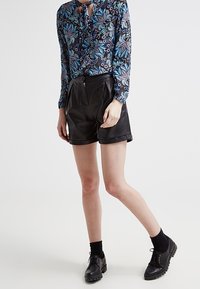 Svarta lädershorts med hög midja och plisserad front, matchade med en blå blus med blommönster som har en nyckelhålsutskärning. Svarta skor och strumpor.