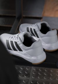 Paire de chaussures de sport blanches avec des rayures noires et des semelles en gomme reposant sur une surface sombre avec un arrière-plan flou.