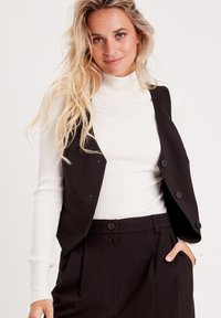 Gilet senza maniche nero con chiusure a bottone sopra un collo alto a coste bianco. Il tessuto appare liscio e ben rifinito, accentuando la silhouette.