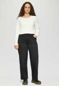 Weißes Langarmoberteil kombiniert mit hochgeschnittenen schwarzen weiten Jeans. Hervorhebenswerte Details sind seitliche Taschen und klobige schwarze Stiefel.