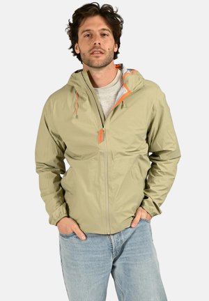 Homme à la barbe portant une veste à capuche beige clair avec une tirette de fermeture éclair orange et un jean bleu clair, debout les mains dans les poches.