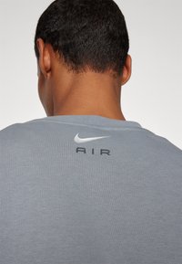 Šedé sportovní tričko s kulatým výstřihem, zdobené stříbrným logem Nike swoosh a nápisem "AIR" v černé barvě, umístěné na horní části zad.
