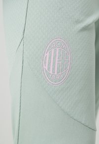 Lichtgroene sportbroeken van textuurstof, met een roze circulair logo met "ACM," "1899," en verticale strepen.