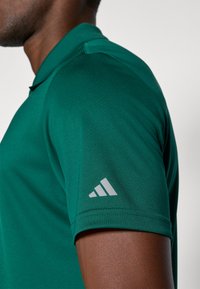 Zelená polokošeľa s hladkou textúrou a priliehavým dizajnom. Obsahuje svetlošedý trojpruhový logo na hornej časti rukáva.