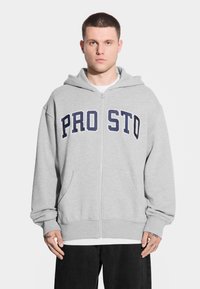 Młody mężczyzna z krótkimi włosami, ubrany w szary rozpinany hoodie z napisem "PRO STO", w połączeniu z białą koszulą i czarnymi spodniami, stojący na białym tle.