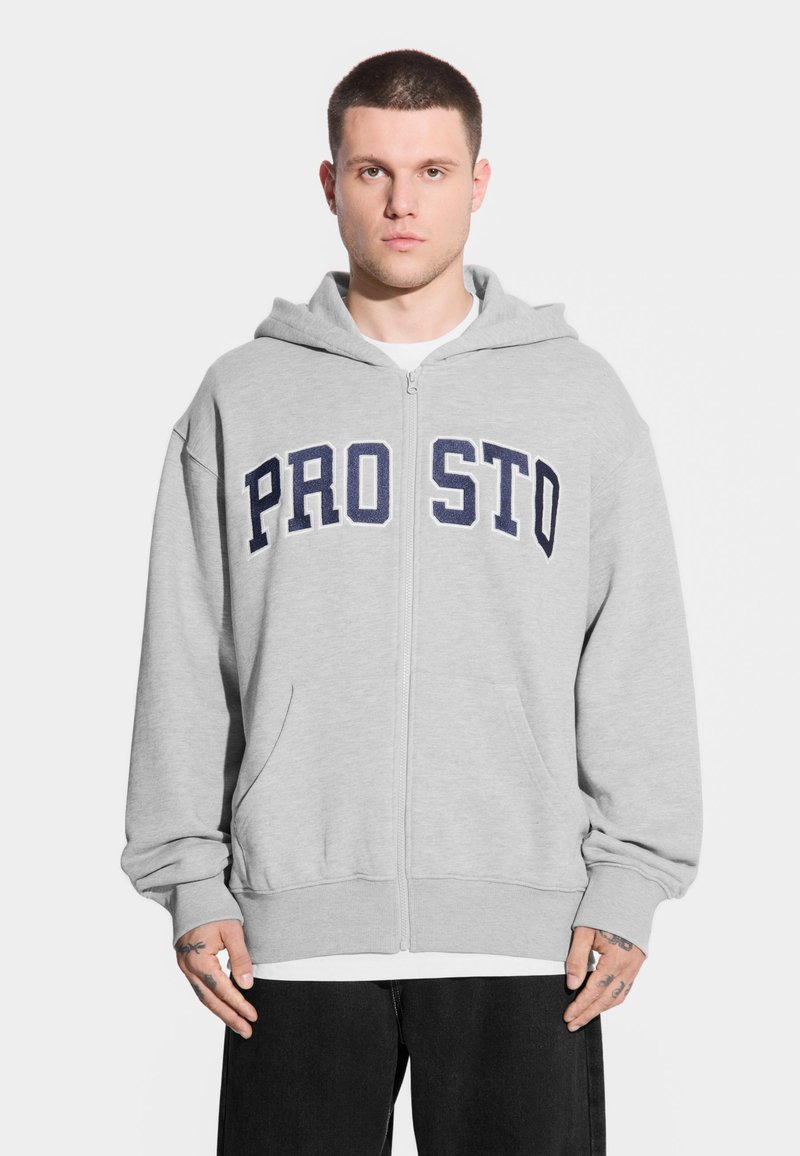 Młody mężczyzna z krótkimi włosami, ubrany w szary rozpinany hoodie z napisem "PRO STO", w połączeniu z białą koszulą i czarnymi spodniami, stojący na białym tle.