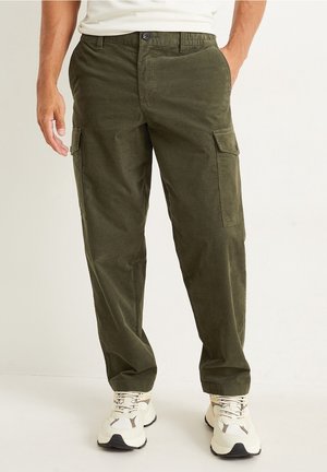 Hombre vistiendo pantalones cargo verde oliva con bolsillos con solapa, combinados con zapatillas blancas, de pie contra un fondo liso.