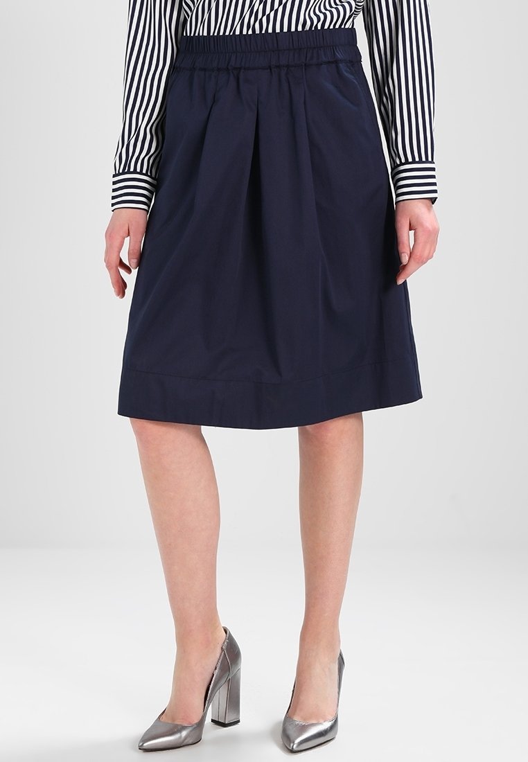 Femme portant une jupe trapèze bleu marine, un chemisier rayé à manches longues et des chaussures à talons hauts argentées métalliques, debout sur un fond blanc.