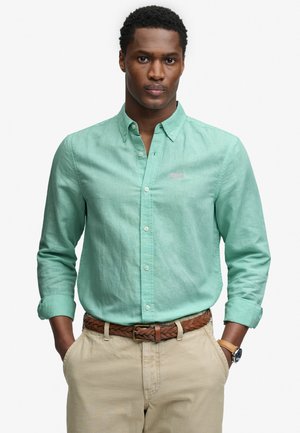 Mężczyzna ubrany w jasnozieloną koszulę z długim rękawem i guzikami, beżowe spodnie, brązowy pleciony pasek oraz zegarek na nadgarstku, stojący z rękami w kieszeniach.